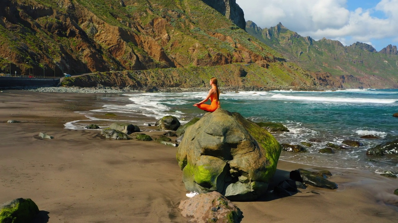 一个成年女孩闭着眼睛在蓝色海水附近的岩石海滩上冥想。女人坐在海边的石头上缓解压力，放松享受平静。视频下载