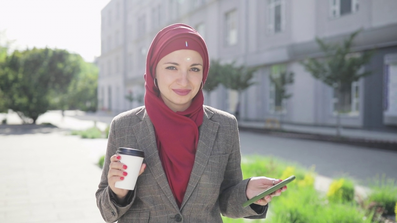 年轻的穆斯林妇女戴着头巾走在市中心，用智能手机喝着咖啡。沟通，网上购物，社交网络概念视频下载