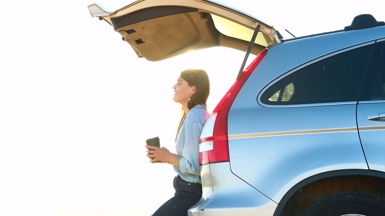 快乐的年轻女子拿着咖啡杯，坐在她的汽车开放的后备箱在麦田日落视频下载
