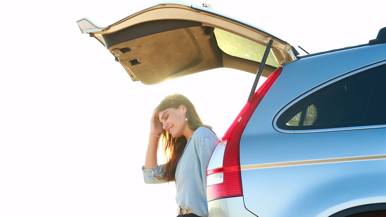 迷人的年轻女子在蓝色衬衫在日落光坐在她的车的开放后备箱视频下载