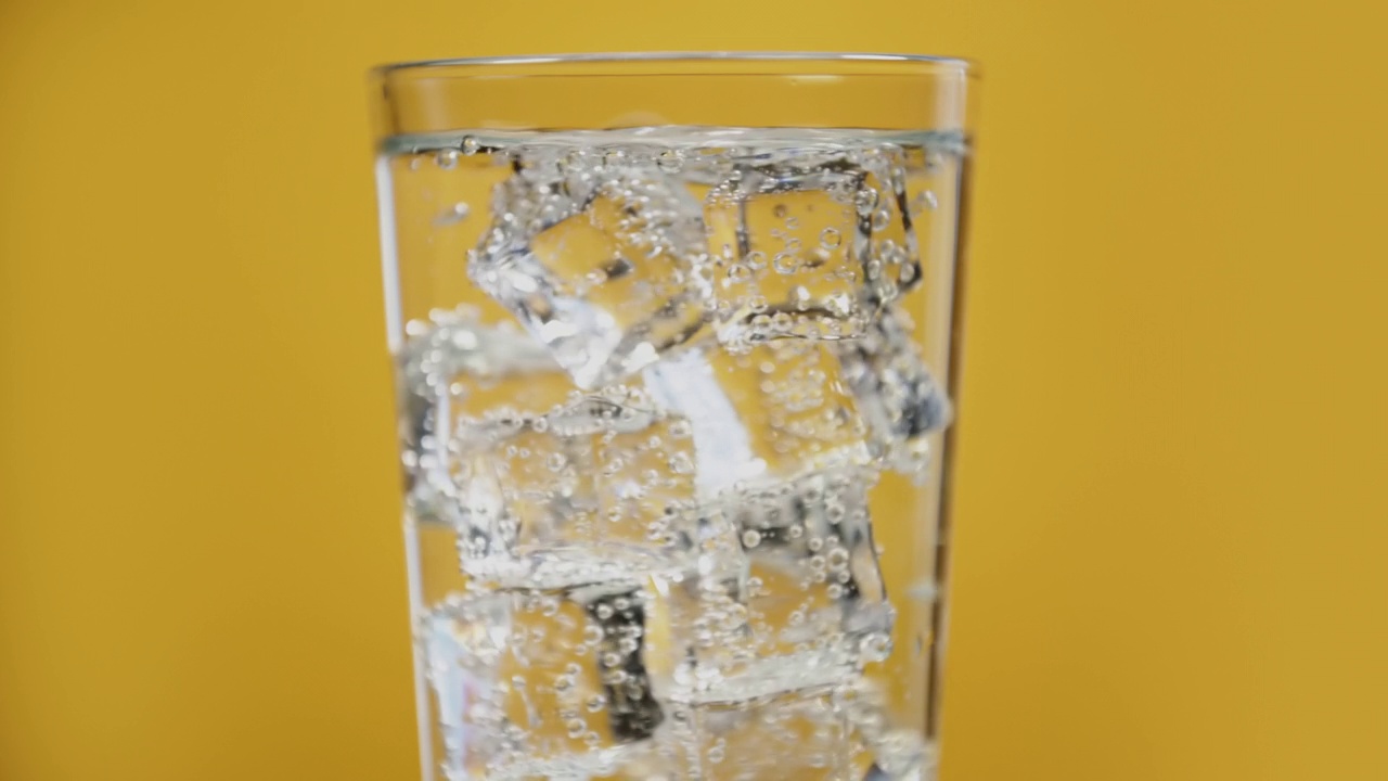 透明碳酸饮料。杯子里装满了冰凉的气泡水，黄色背景上有冰块视频素材