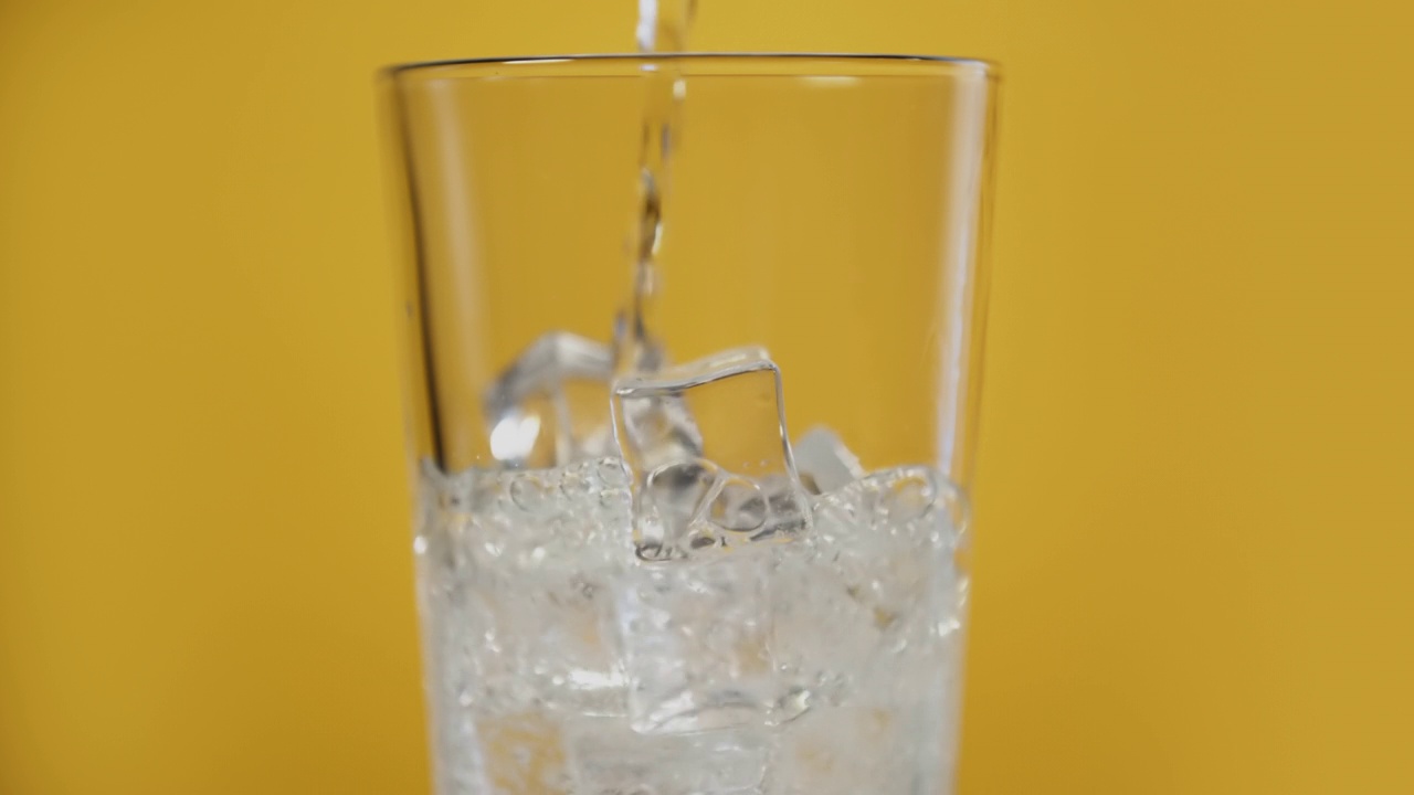 在玻璃杯中倒入透明的碳酸饮料，黄色背景上有冰块视频素材