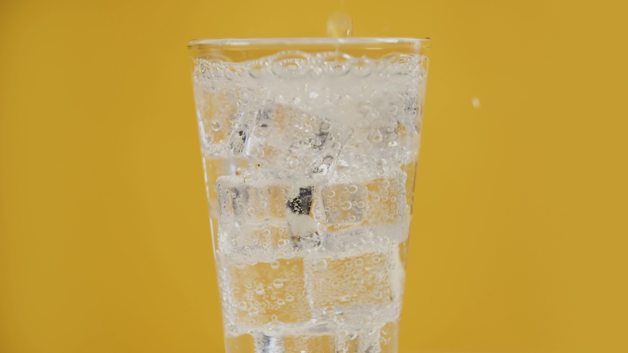 在玻璃杯中倒入透明的碳酸饮料，黄色背景上有冰块视频素材