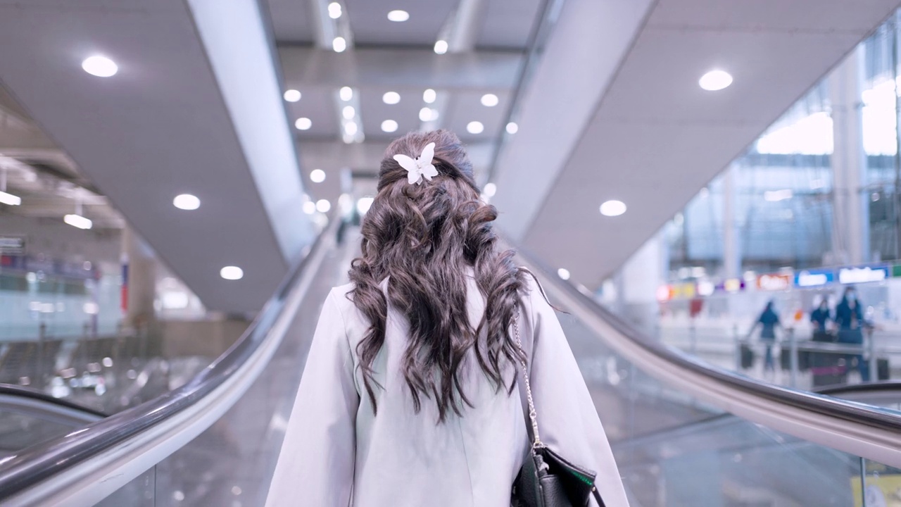 亚洲女商人戴着口罩站在机场航站楼的自动扶梯上的后视图视频素材