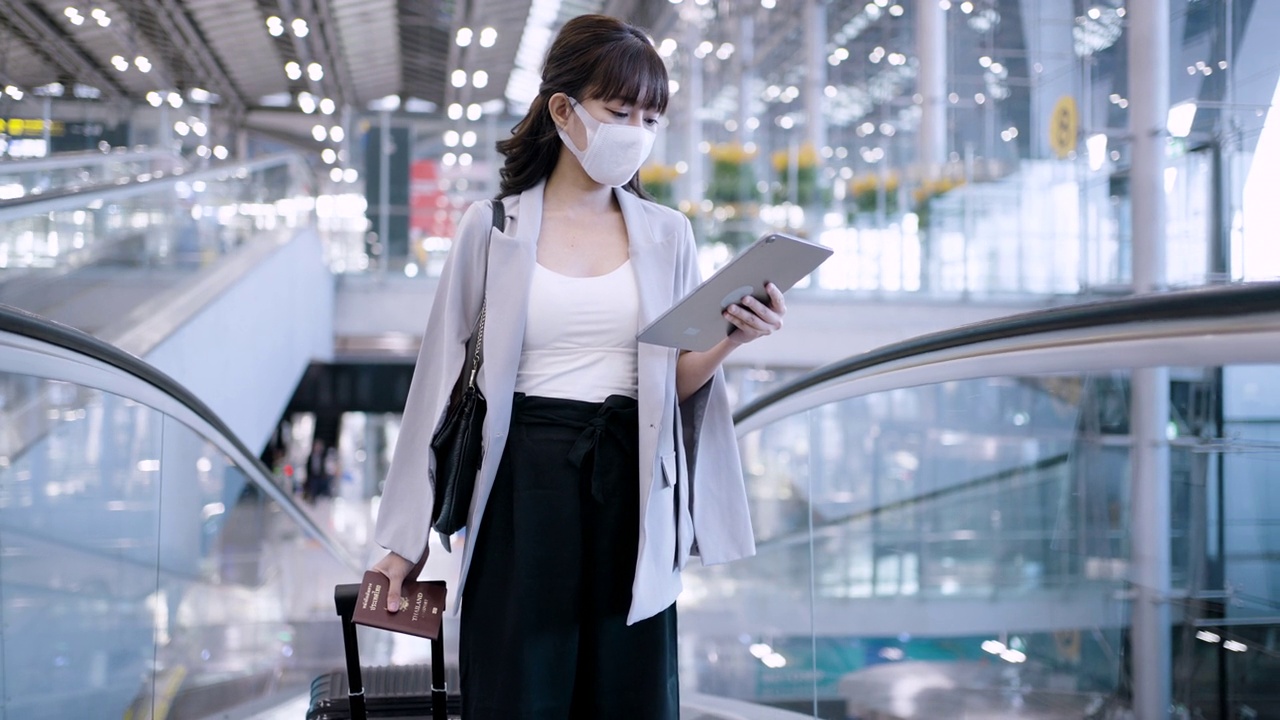 一名亚洲年轻女商人戴着口罩，在机场航站楼的自动扶梯上使用平板电脑视频素材