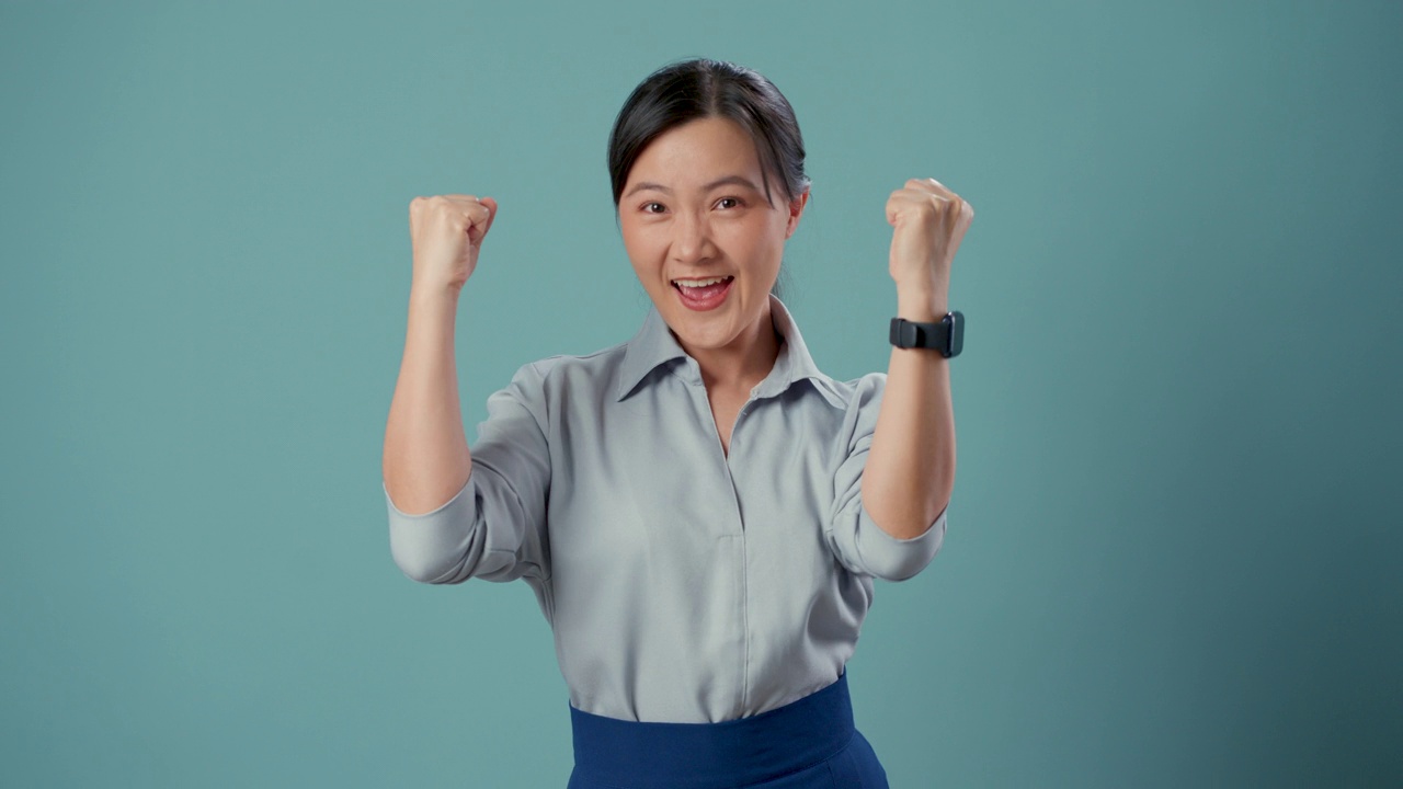 亚洲妇女高兴自信地展示她的拳头，使一个胜利的手势站在蓝色背景孤立。视频购买