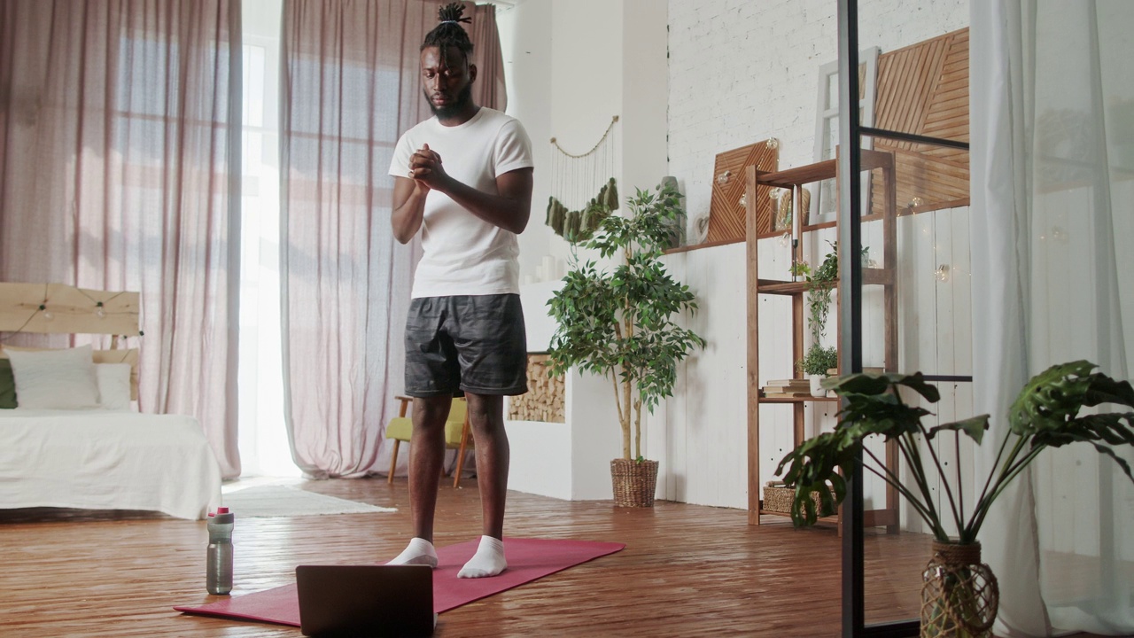 年轻的初学者非洲裔美国人在客厅的地板上做体操练习。有吸引力的帅气运动男性花休闲活动时间锻炼保健通过在家里的笔记本电脑上的在线指导视频下载