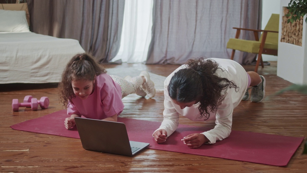 妈妈和可爱的小女儿一起做平板支撑练习，在舒适的家里打开笔记本电脑。家庭保健和运动心理治疗在家里的概念。视频下载