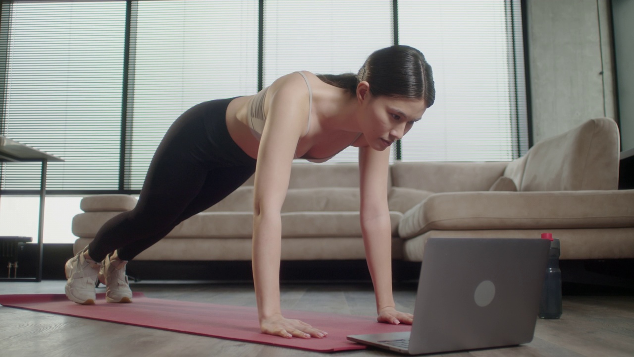运动的亚洲妇女在运动服工作在家。健身女在打开的笔记本电脑前摇着瑜伽垫，重复着专业健身教练观看在线视频教程的指导视频下载