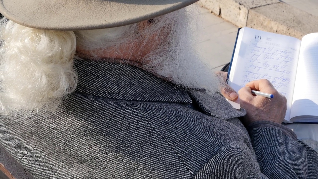 俯视后面的老人，灰色胡子和白色辫子，头上戴着帽子，穿着灰色外套，坐在户外的长凳上，用钢笔在笔记本上写笔记视频下载
