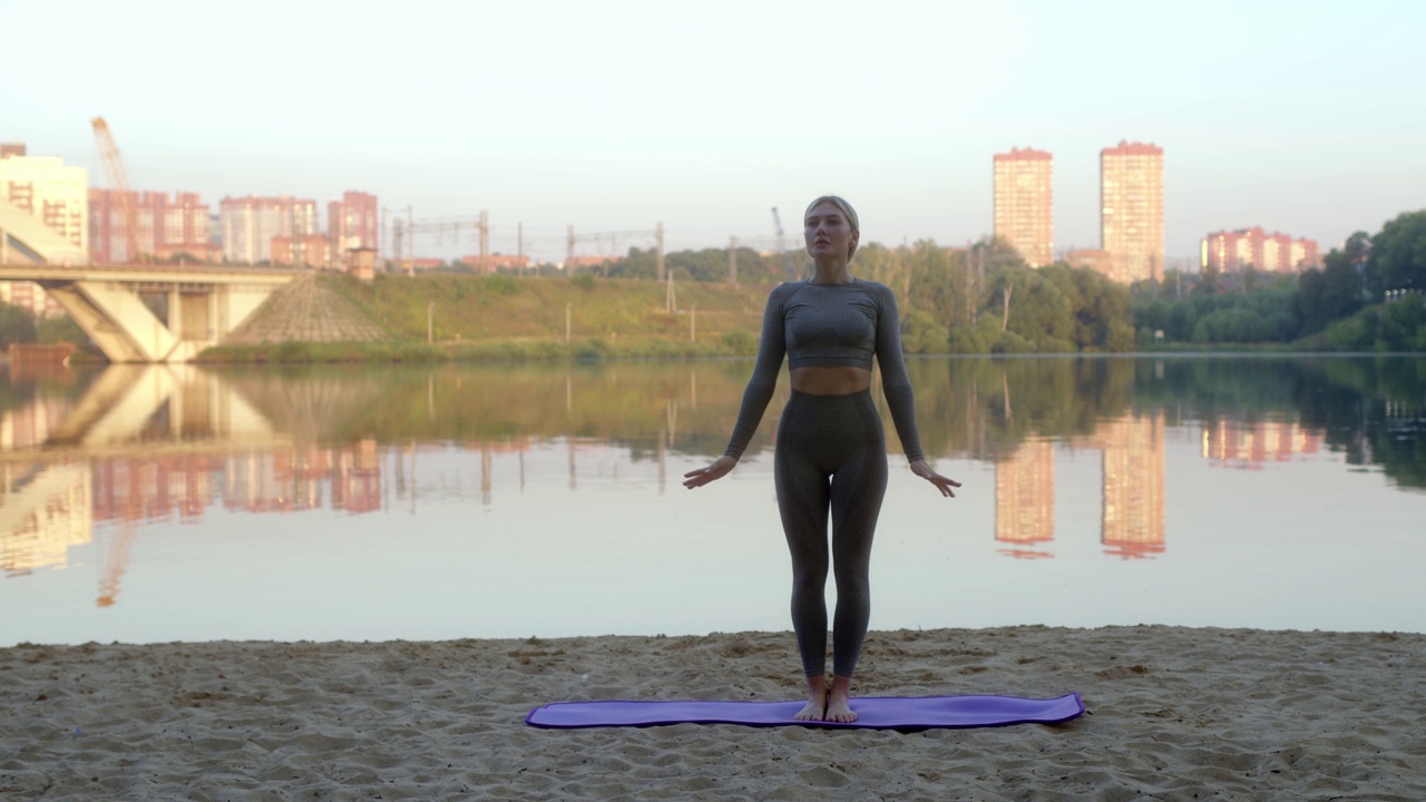 性感苗条的年轻女子与金发马尾做早上伸展在河岸灰色运动服在紫色运动垫。漂亮的女孩热身，做瑜伽体式。视频素材