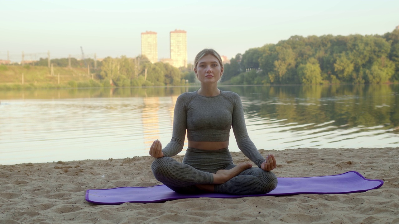 身穿灰色紧身运动服的年轻女子坐在河边或湖边的紫色沙滩垫子上，看着镜头。放松放松，事业有成视频素材