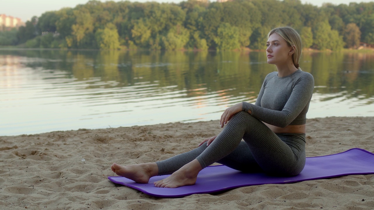 苗条的年轻女子坐在紫色的运动垫在沙滩岸边的河，看着远方。身穿灰色紧身衣的健康女孩在运动后休息。思考世界上正在发生的事情。视频素材