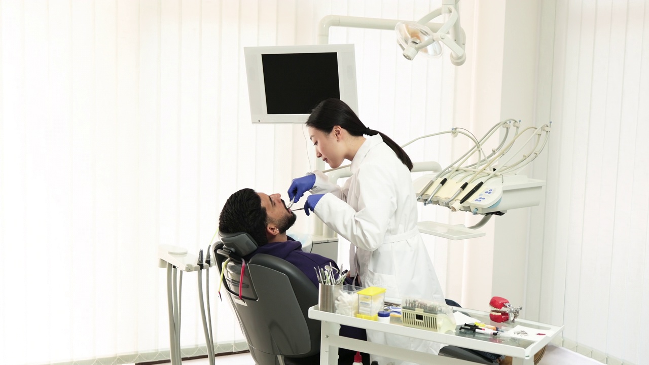 亚洲牙医正在用牙钻治疗一名年轻胡须男子的牙齿视频下载