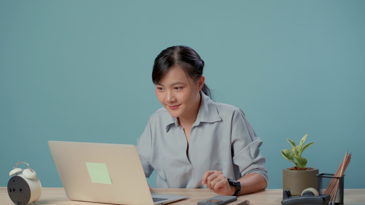 亚洲妇女把食指放在嘴唇上，坐在办公室里，被蓝色背景隔离。视频素材