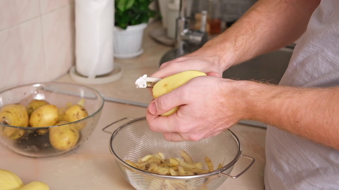 在明亮的厨房里，一名男子用蔬菜削皮机剥土豆皮。煮土豆的准备工作视频素材