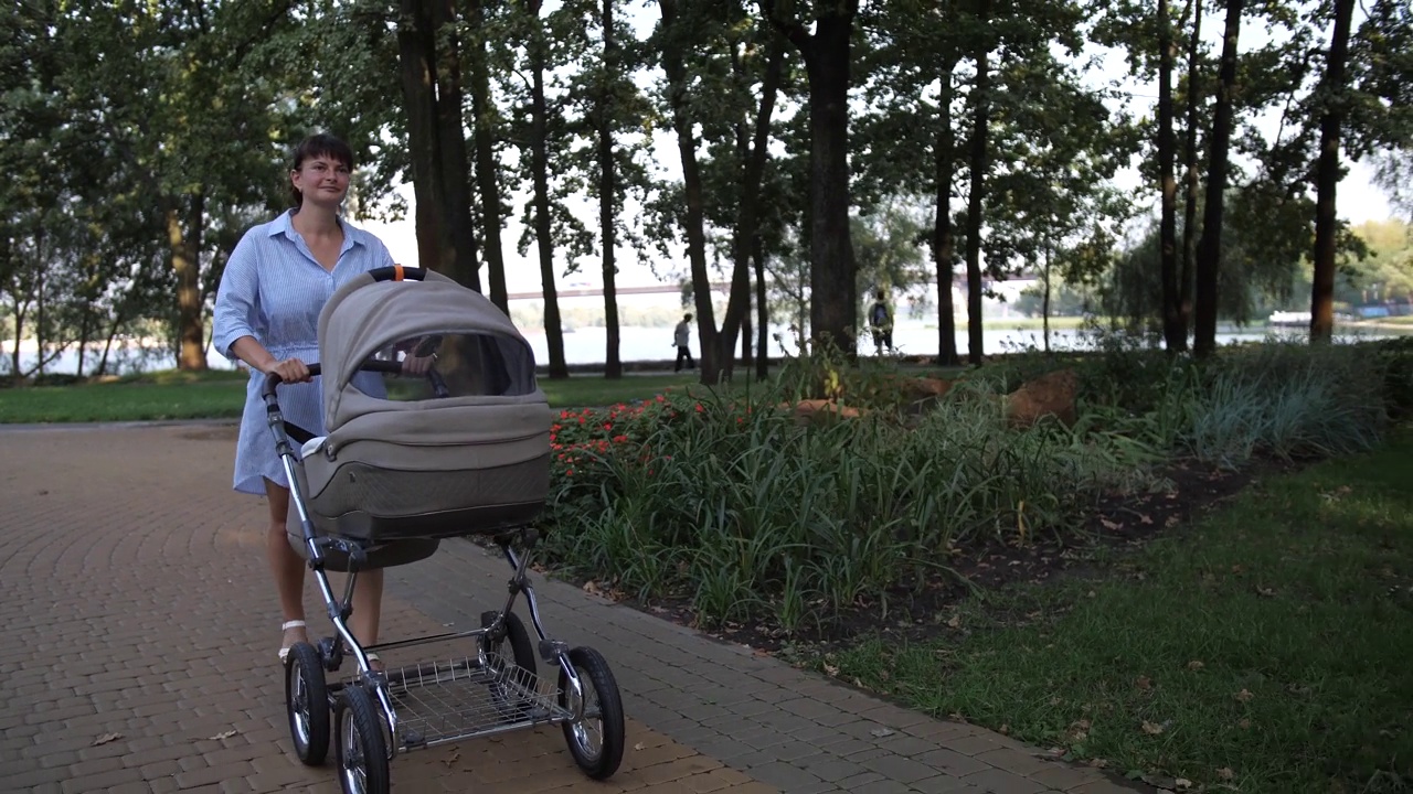 一位年轻的母亲推着婴儿车在公园里散步。视频下载