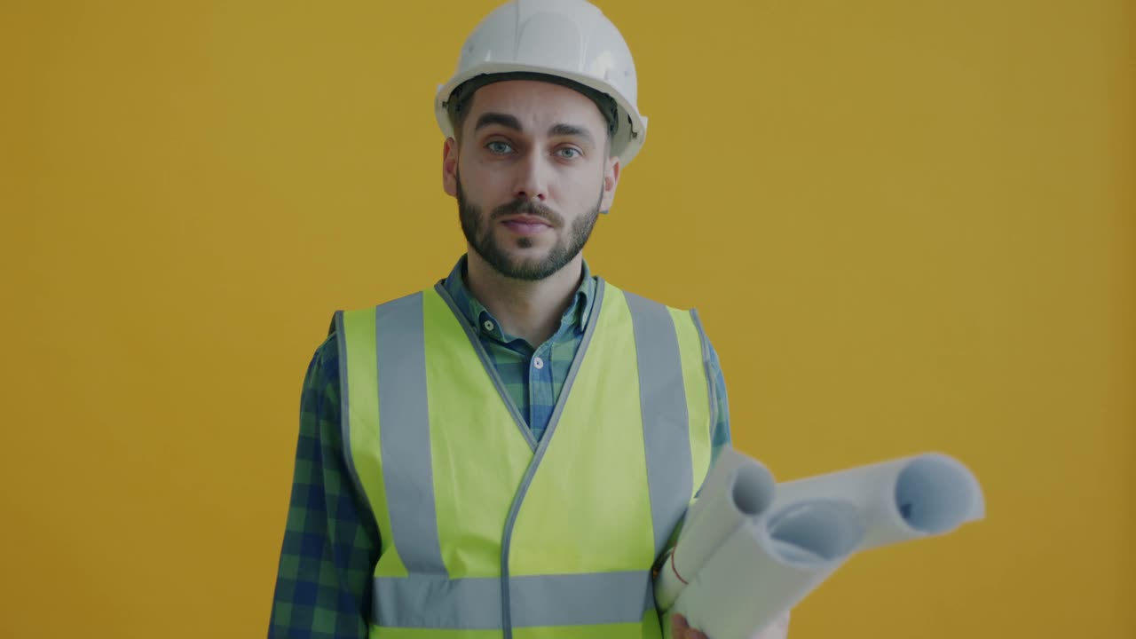 慢动作肖像工业工程师拿着蓝图触摸头盔和微笑在黄色背景视频素材