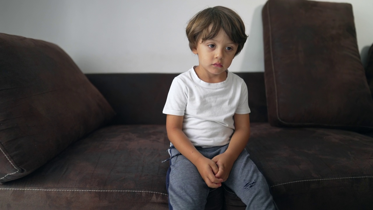 一个无聊的孩子坐在家里的沙发上无所事事。年轻的男孩带着沉思的表情感到无聊视频素材