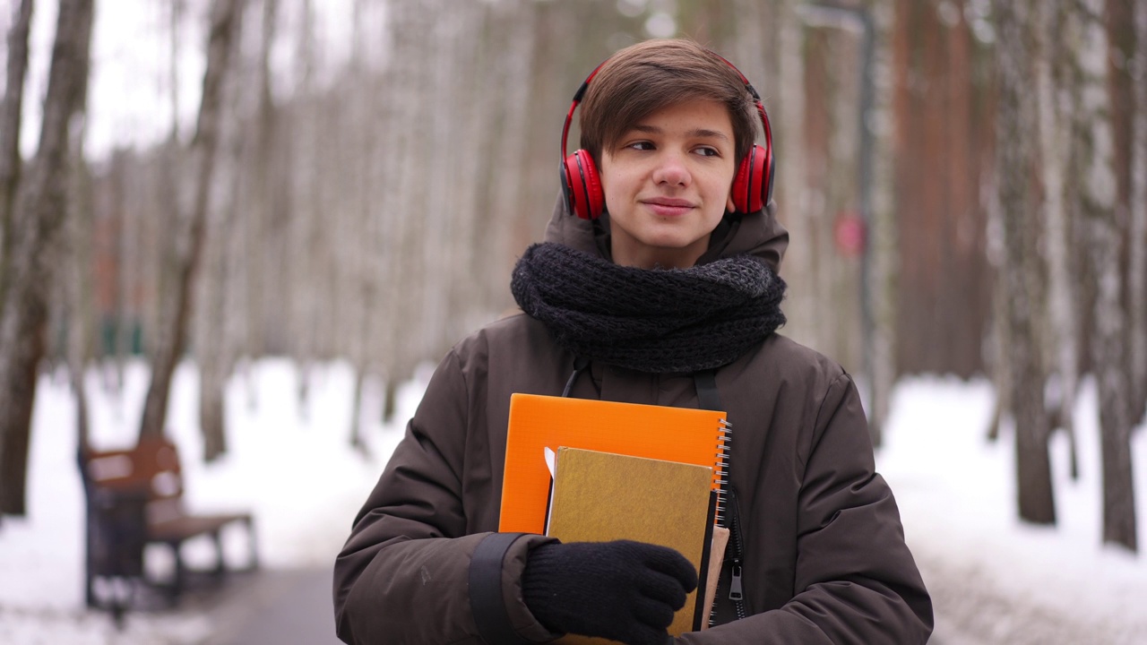 自信聪明的少年戴着耳机在公园里学习后散步，听着音乐微笑着离开。肖像的快乐满意的白人少年在户外漫步慢动作。视频下载