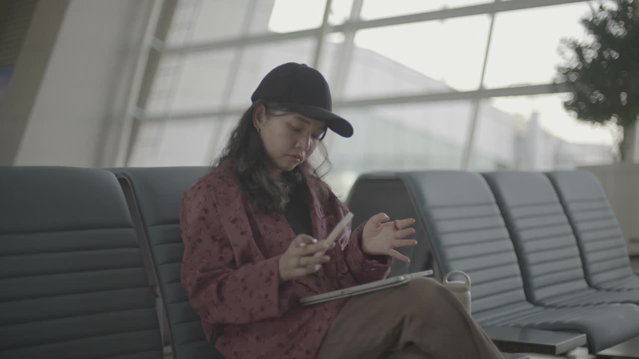 女商人坐在登机口附近的机场候机区用平板电脑和智能手机工作。她边喝水边环顾四周。视频下载