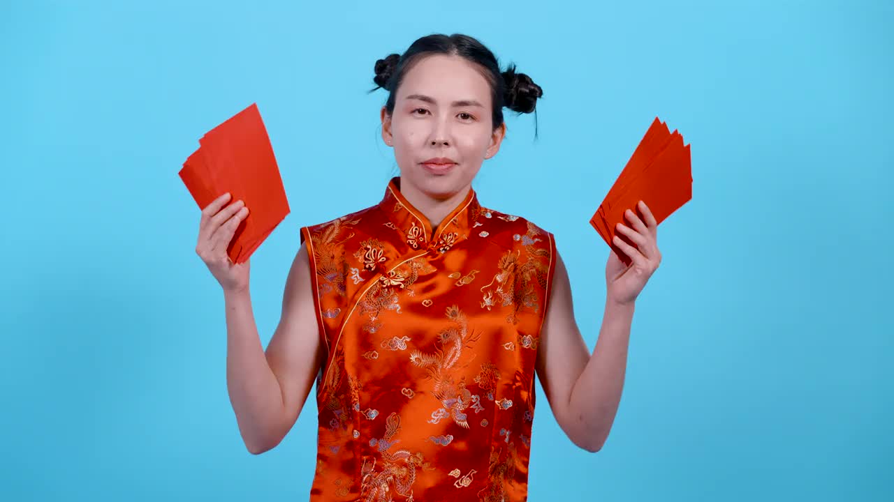 4K，肖像，女孩在中国新年穿着旗袍，他的面部表情，非常高兴和高兴，收到许多红包，手满，孤立的室内工作室在蓝色背景。视频下载