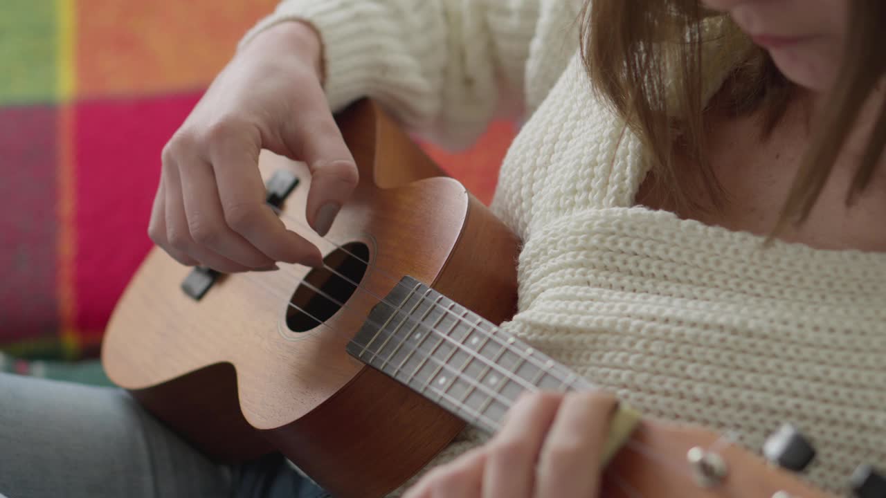 女性手中的尤克里里。一个女人坐在家里的沙发上学习弹奏尤克里里。这个女孩用一把小吉他弹着尤克里里，然后唱歌。爱好和音乐弦乐器概念。视频下载