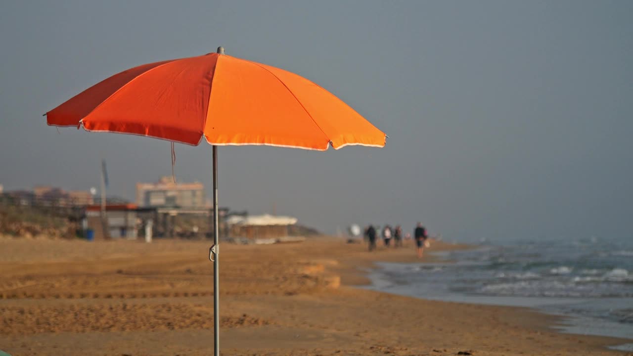 一张橙色的沙滩伞，背景是海滩上的行人，用慢动作拍摄。背景是海滩酒吧和建筑物。视频下载