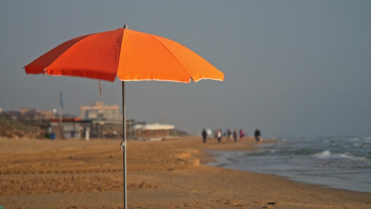 一张橙色的沙滩伞，背景是海滩上的行人，用慢动作拍摄。背景是酒吧和建筑物。视频下载