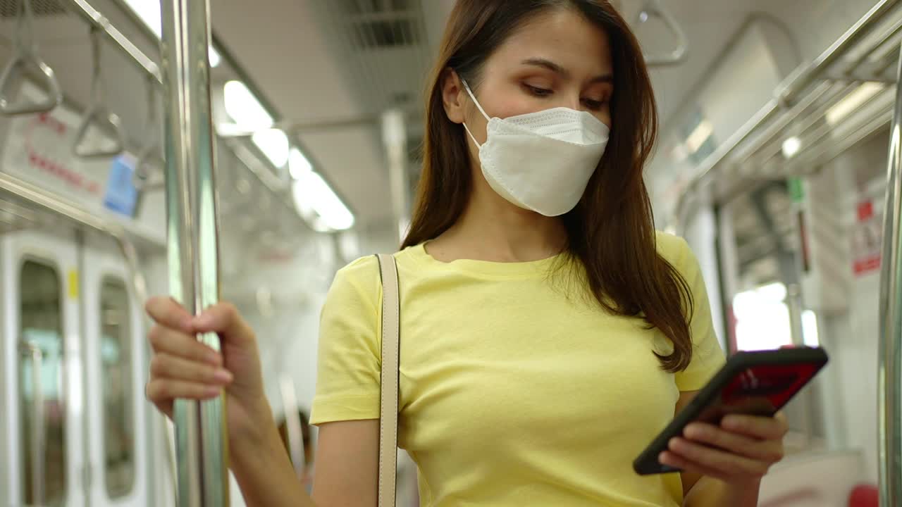 年轻女性旅行者佩戴预防疫情暴发和城市的防护口罩，在城市公共交通工具上使用手机视频下载