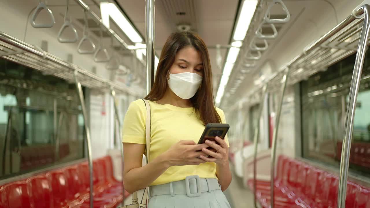 年轻女性旅行者佩戴预防疫情暴发和城市的防护口罩，在城市公共交通工具上使用手机视频下载