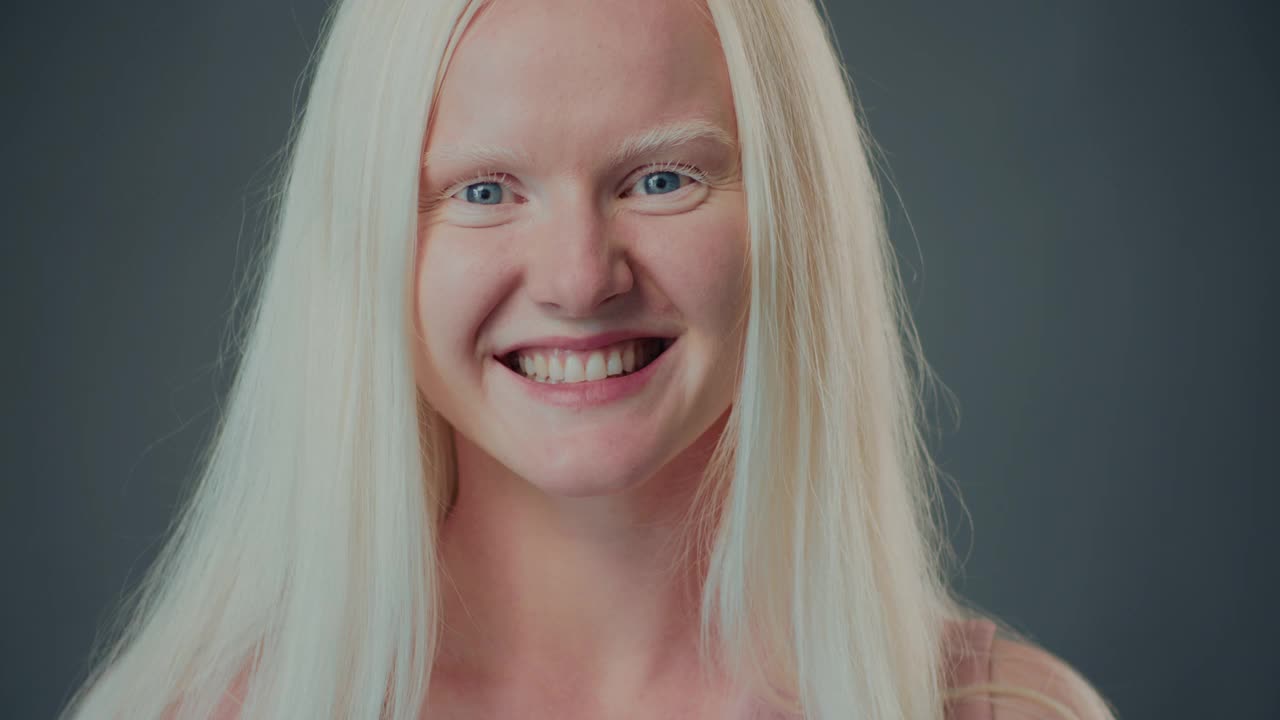 特写肖像美丽快乐的年轻20岁白化病妇女触摸头发微笑在相机在灰色背景。视频下载