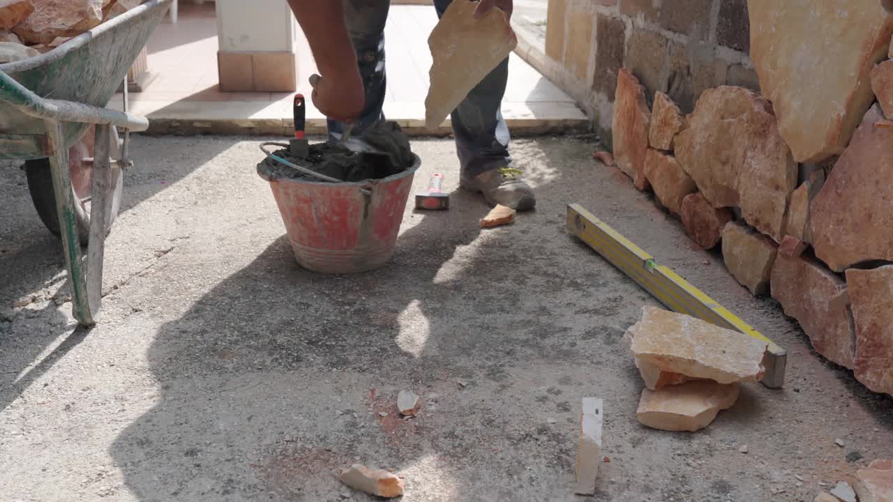 砖瓦工用泥铲将混凝土溶液涂在岩石面粘贴在墙面上进行装饰，建筑工在施工现场进行石工，用不同大小形状的橘色砖进行墙面装饰视频素材