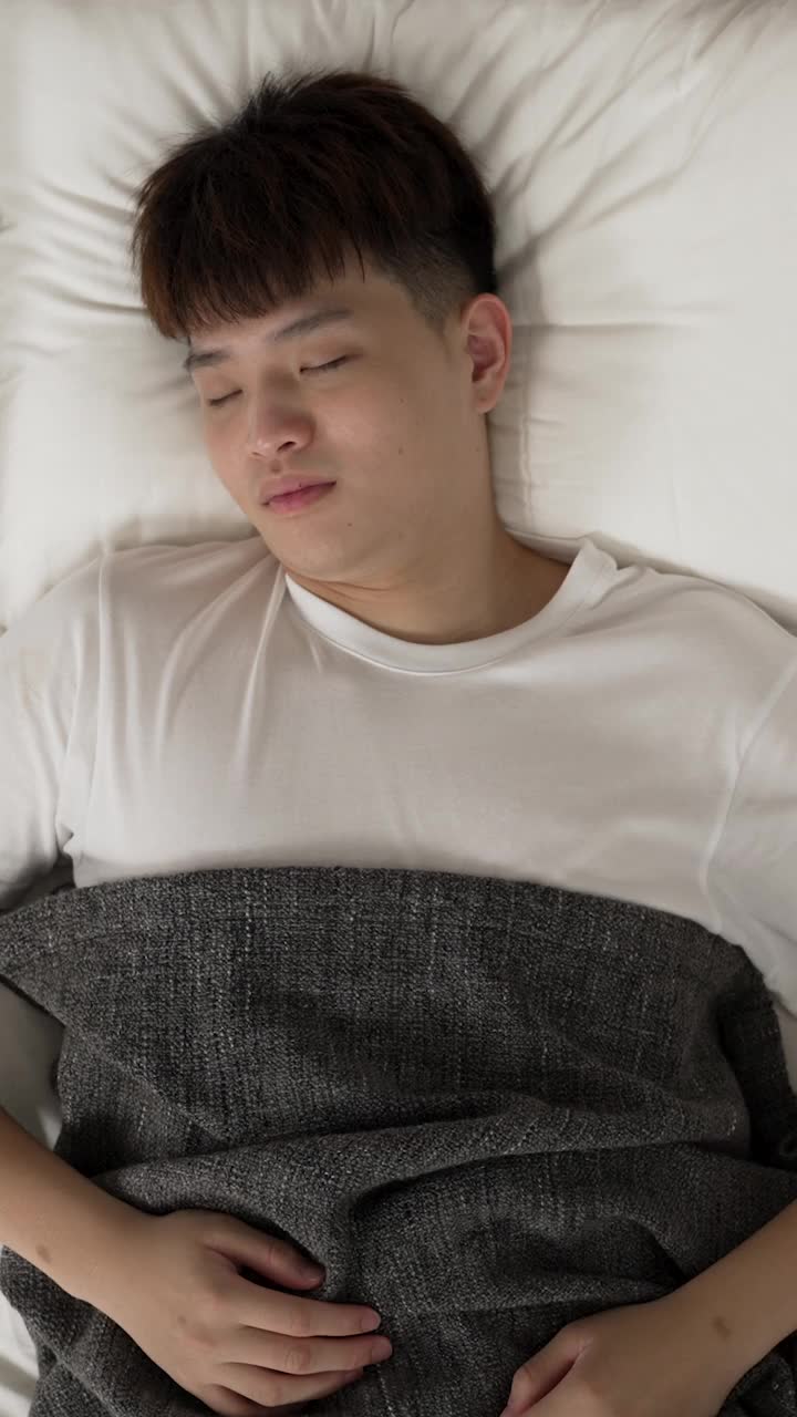 垂直屏幕:一个亚洲男人在床上翻来覆去，早上几乎从睡眠中醒来，在一个现代家庭卧室的顶部视角。视频素材