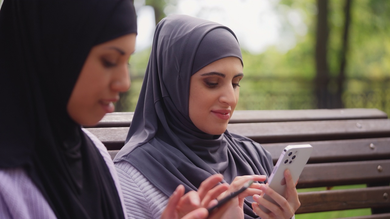 两名多民族穆斯林妇女在公园里滚动智能手机，使用社交媒体应用程序视频下载