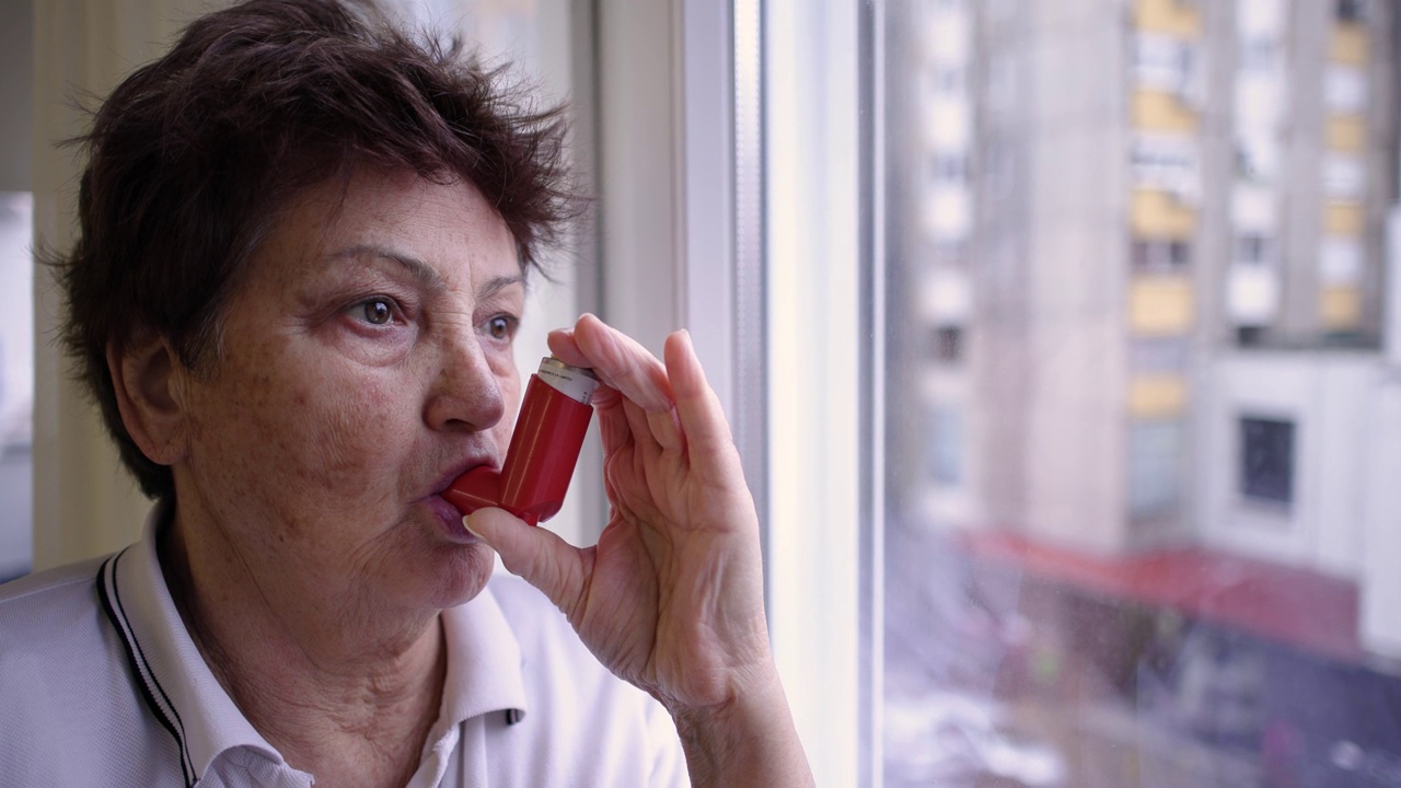 老年妇女一边用哮喘吸入器，一边透过窗户往外看视频下载