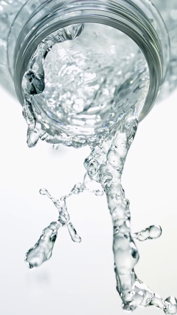 特写和超慢镜头的瞬间，大量晶莹剔透的水从倾斜的透明水瓶壶嘴中喷涌而出，在一个垂直的白色背景上视频素材