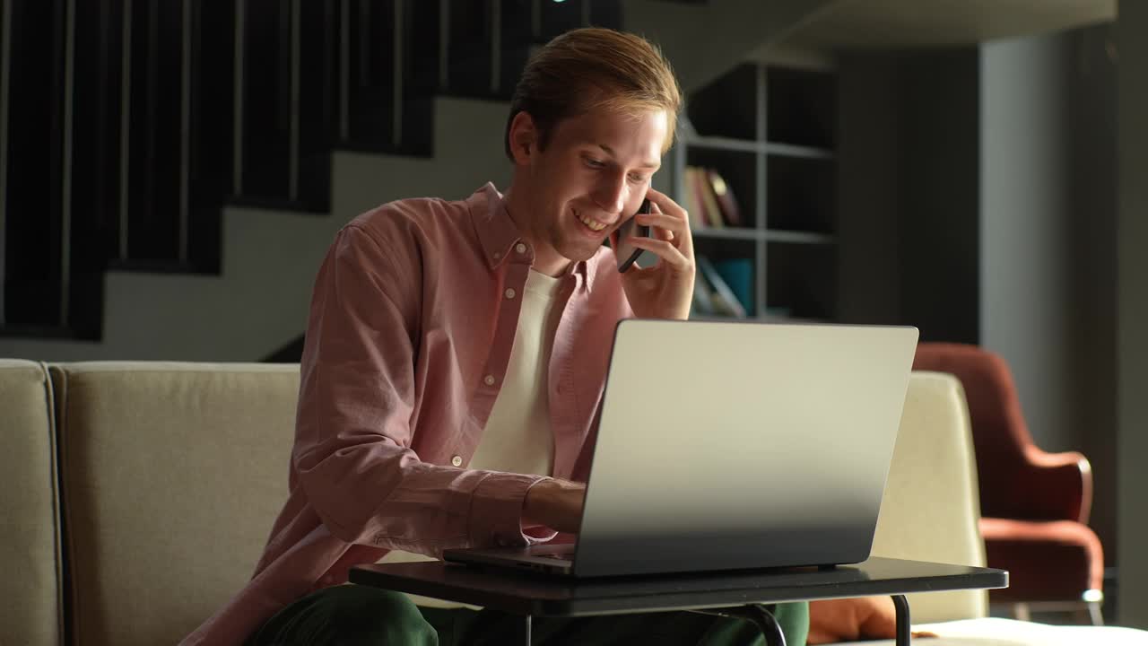 开朗的年轻白人男子坐在沙发上的办公桌，在笔记本电脑上工作，在家里讲手机。积极的男性自由职业者或计算机专业学生视频素材