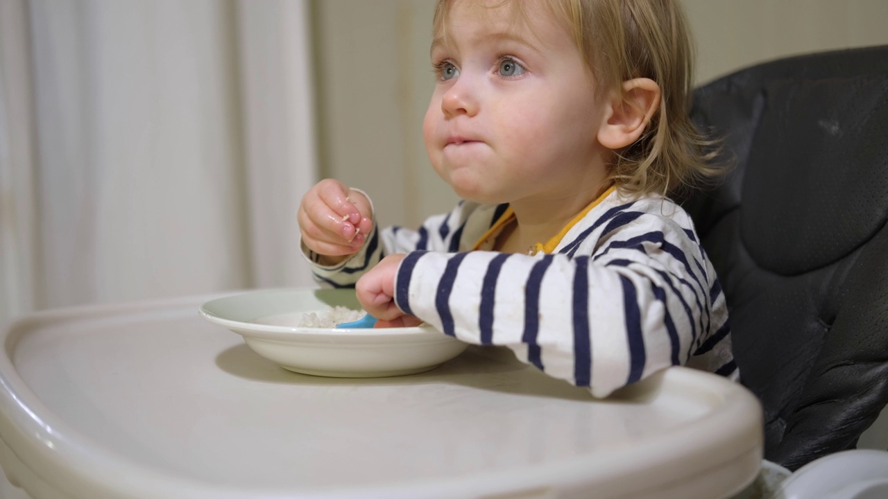 一名一岁的饥饿女婴，穿着条纹休闲服，坐在灰色高脚椅上的白色桌子旁，自己用勺子吃米饭。模糊的餐厅背景。孩子的健康饮食。孩子的营养。视频素材