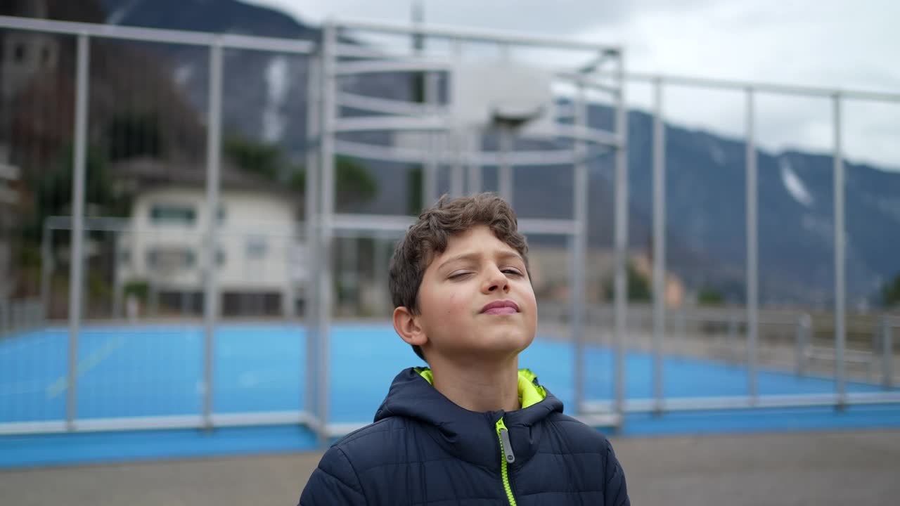 一个英俊的小男孩站在外面，表情严肃地看着相机。一个十几岁的孩子站在城市公园的户外。跟踪拍摄视频素材