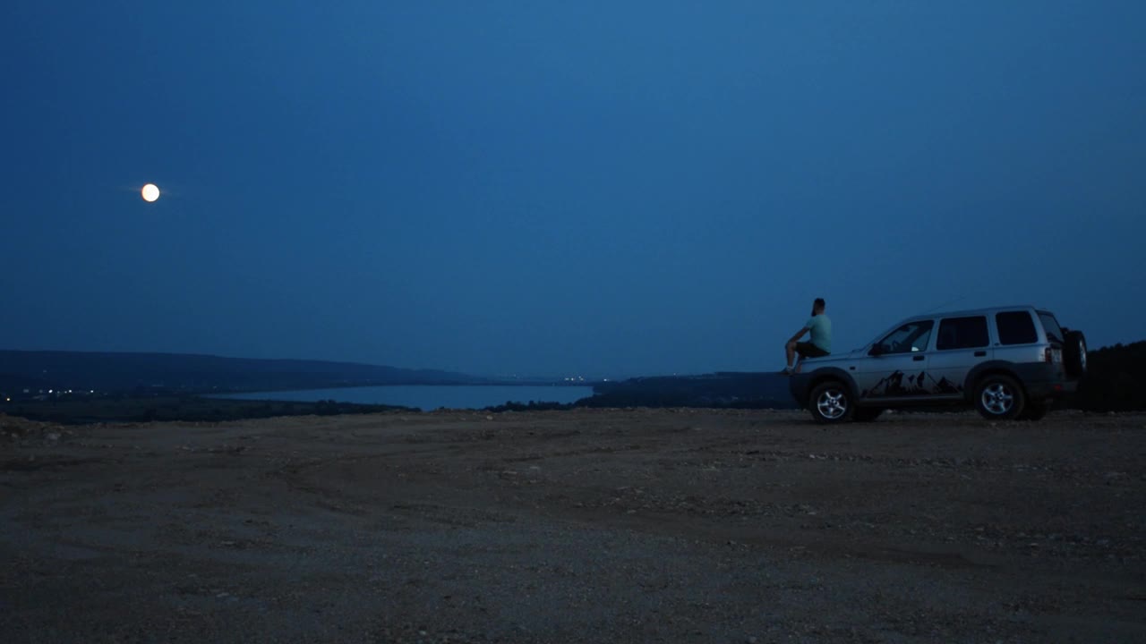 一个男人独自坐在河边的汽车引擎盖上，在夜空下凝视着远方视频下载