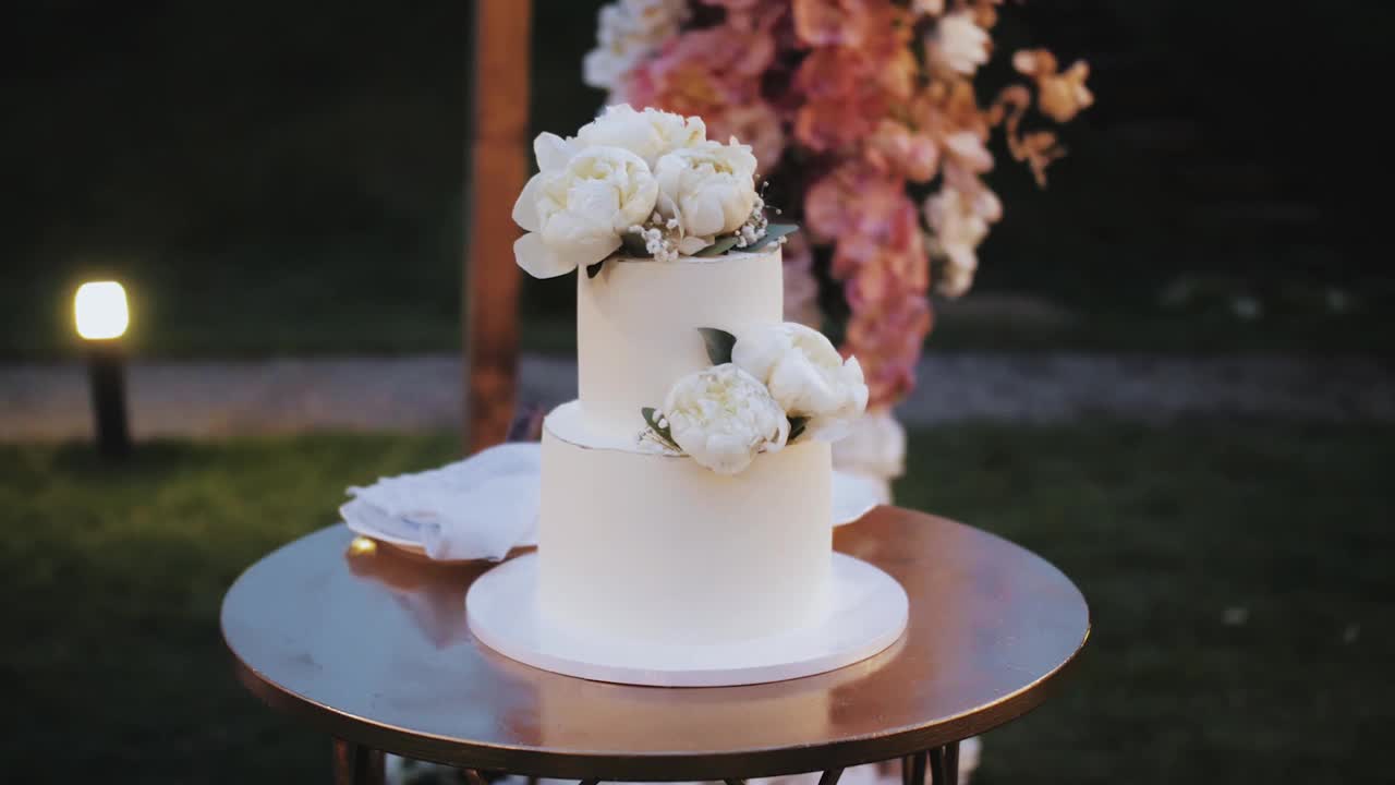 用白色牡丹花装饰的婚礼蛋糕慢动作。美丽的婚礼蛋糕白色，婚礼拱门的背景。视频下载