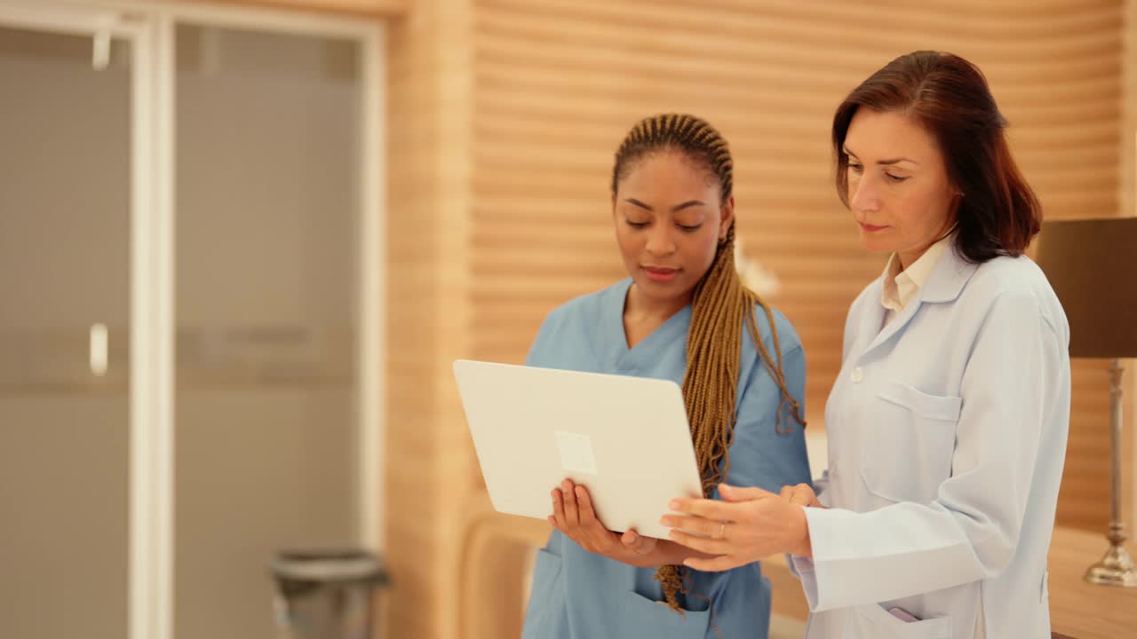 图为，在医院接待处，非洲裔年轻护士正拿着笔记本电脑，向白人女医生咨询患者的病情。视频素材