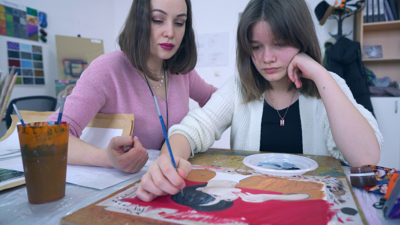 一个十几岁的女孩，一个有抱负的艺术家，用画笔和颜料在纸上画画，一位女老师在提示她。视频下载