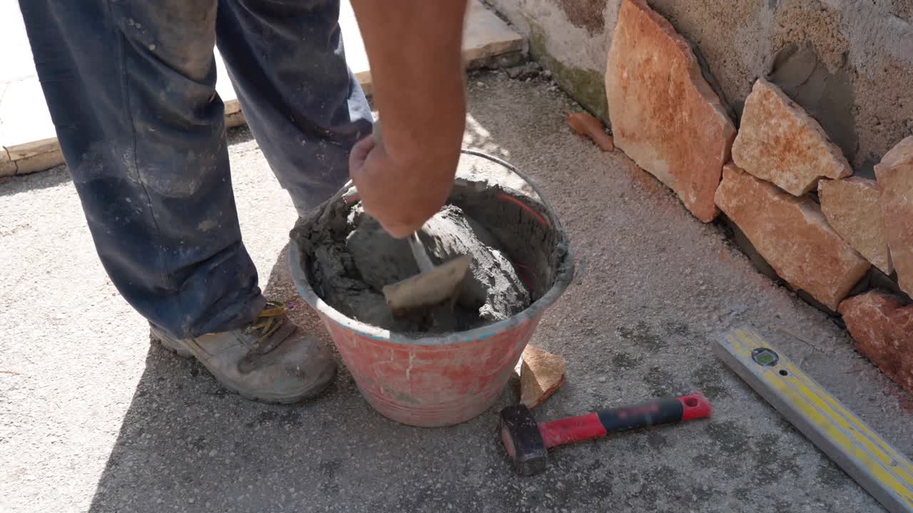 泥瓦工穿着脏衣服，用手铲在桶里搅拌混凝土溶液，制作装饰石材贴墙面用湿水泥，施工现场砌砖视频素材