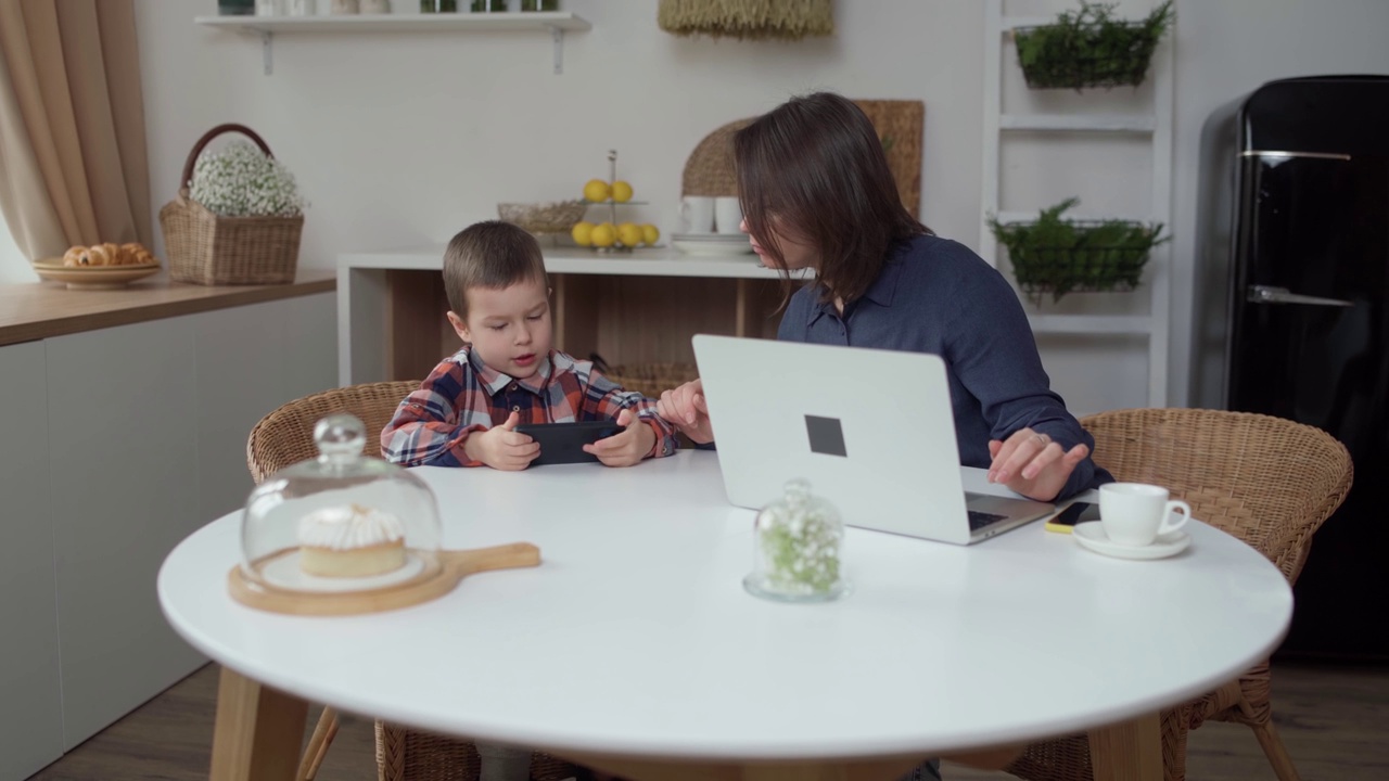 一个女人带着她的小儿子坐在桌子前，用笔记本电脑工作。小孩使用电话视频下载