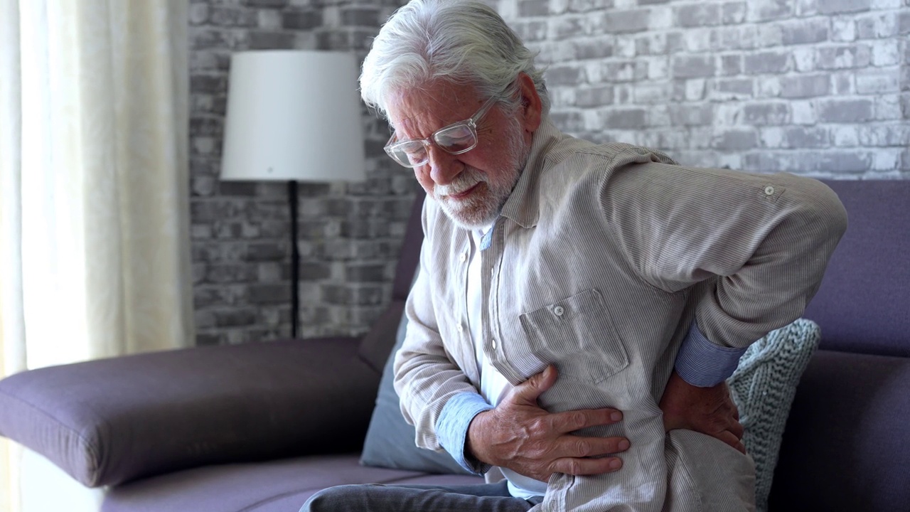 心烦意乱的老年男子感到突然腰酸背痛肌肉酸痛，坐在家里的紧张伤，伤心的老爷爷触摸脊柱有腰痛，腰痛骨关节炎，腰痛的概念视频下载