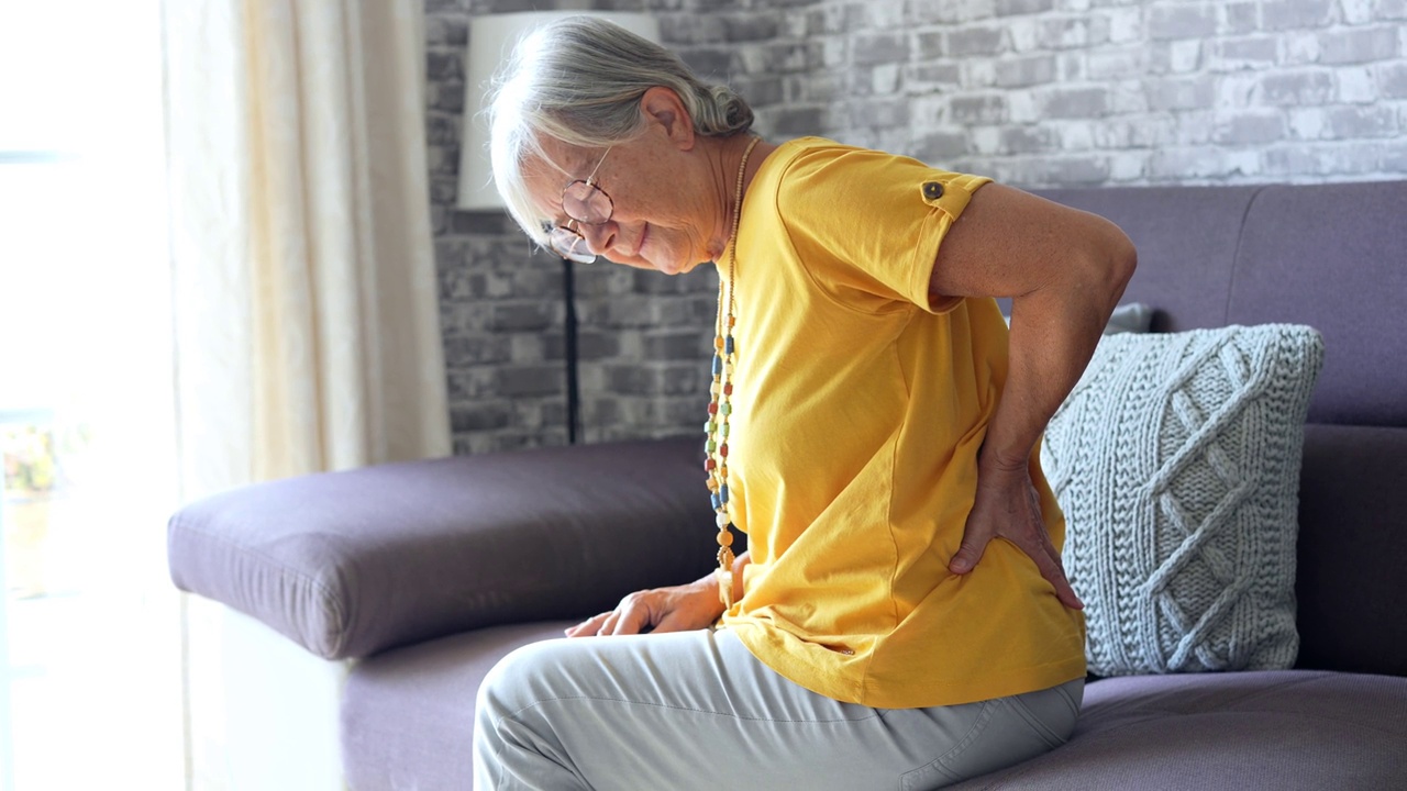不快乐的成熟女性抚摸背部，感觉疼痛，坐在客厅沙发上，不健康的中年老年女性遭受背痛，脊椎问题，摩擦僵硬的肌肉或肾脏视频下载