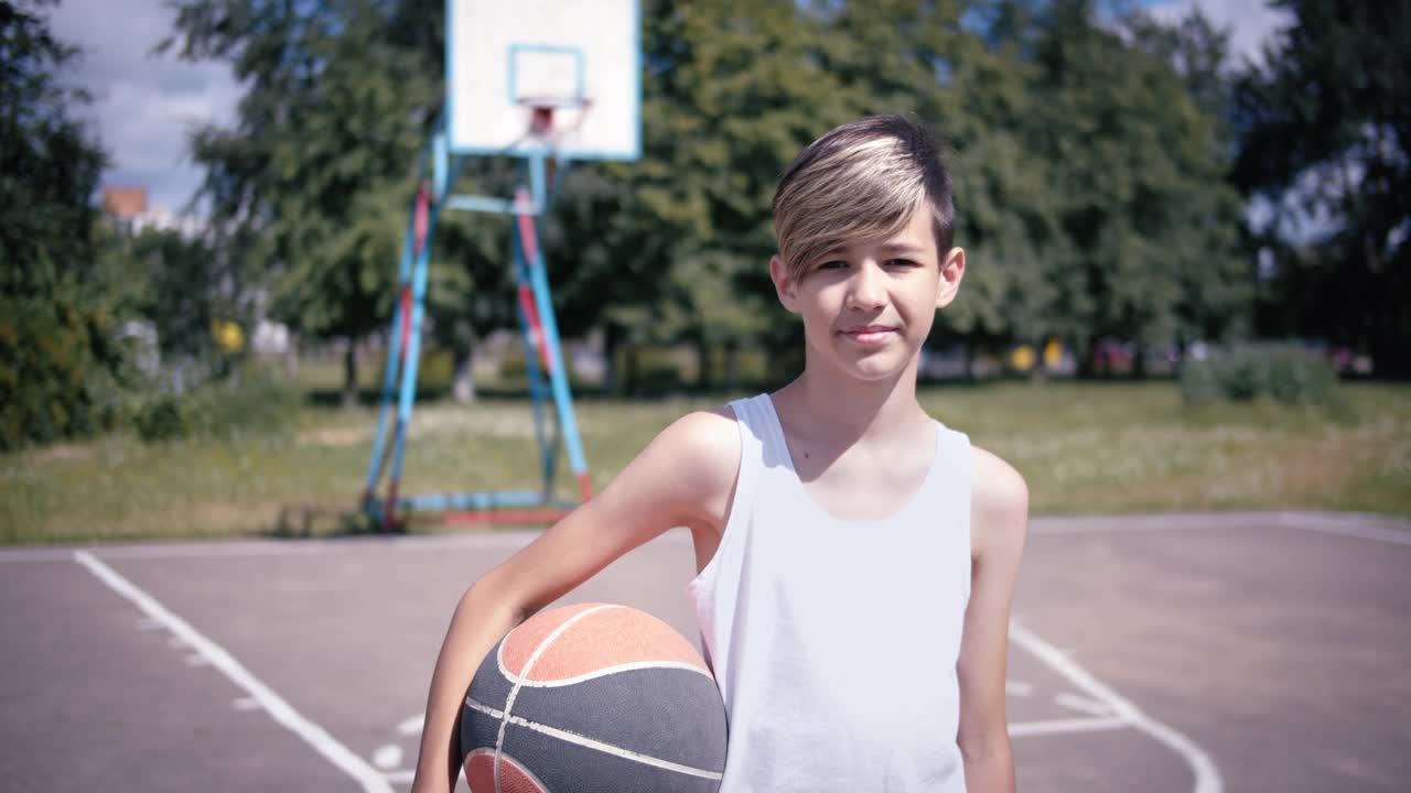 一个男孩在篮球场上拿着球看着镜头微笑，镜头移动，定格拍摄视频下载