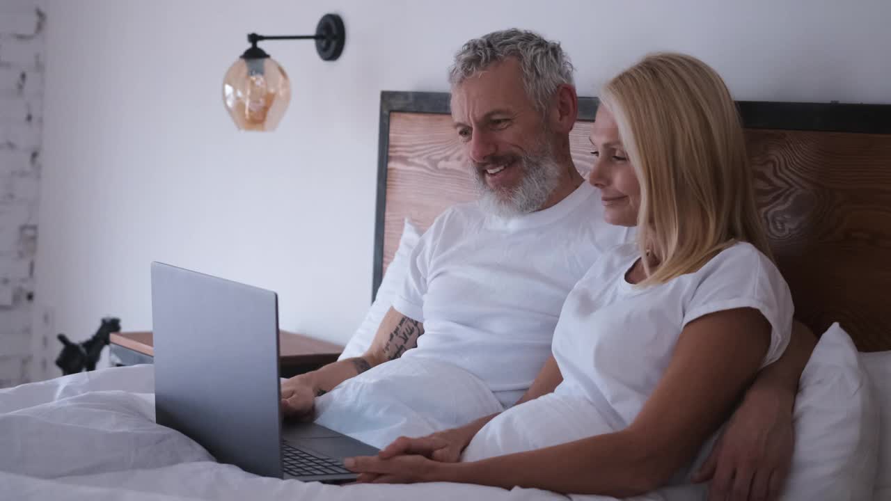 那对幸福的夫妇坐在床上玩笔记本电脑。慢动作视频下载